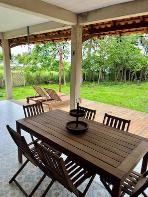 A wooden table with chairs, a hanging light fixture, and a view of a green garden. - #1 Rosário Marajó Residence - Água Boa - One Suite (Salvaterra)