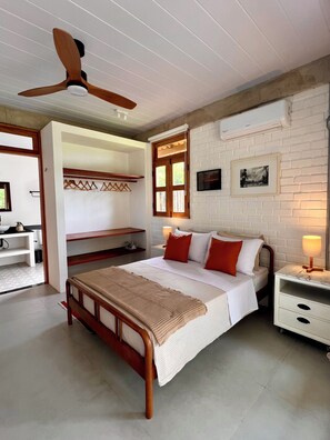 Une chambre avec un lit en bois, un ventilateur de plafond, un mur en briques blanches, une fenêtre avec des volets en bois et une table de chevet avec une lampe.