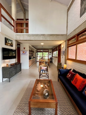 A spacious living area with a high ceiling, a television, a glass coffee table, a sofa with cushions, and a dining area in the background.