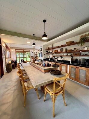 A modern kitchen with a long island, wooden chairs, pendant lights, and open shelving with kitchenware.