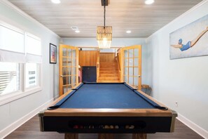 A pool table in a room with wooden flooring, a ceiling light, and a painting on the wall. - 8 Bed Oceanfront w Pool/Spa, Private Beach Access! (Nags Head)