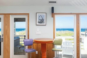 A room with a wooden table and chairs, a framed picture on the wall, and a view of the ocean through large glass doors. - 8 Bed Oceanfront w Pool/Spa, Private Beach Access! (Nags Head)