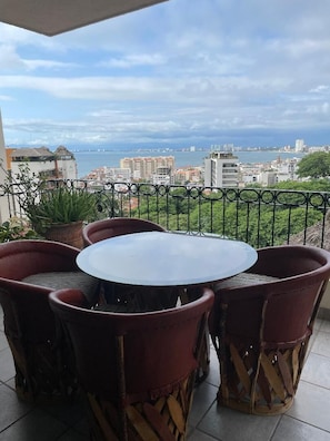 Ein runder Tisch mit sechs Stühlen auf einem Balkon mit Metallgeländer, Blick auf Stadt und Meer.