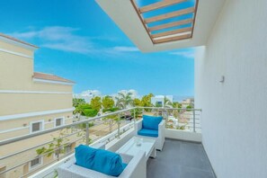 A balcony with a view of buildings and greenery, a white wall, and a metal railing.