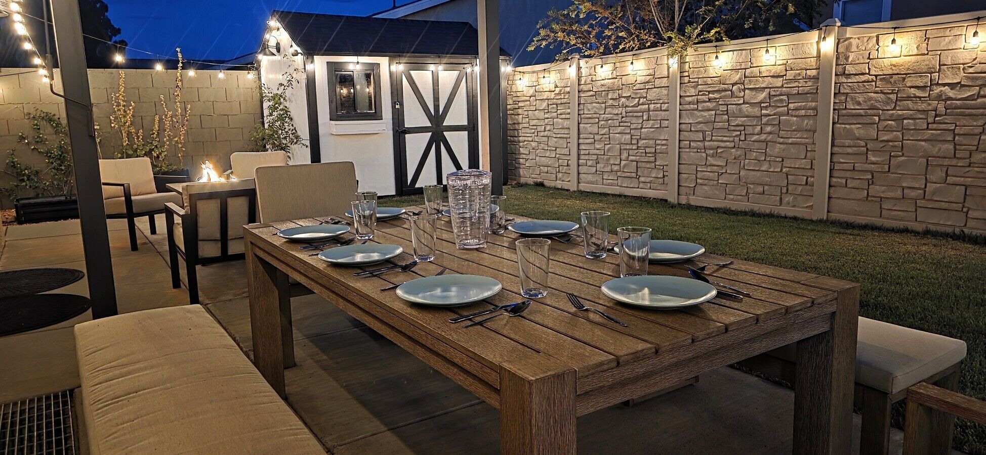Een buiten eethoek met een houten tafel gedekt voor zes personen, omringd door stoelen en een stenen muur.