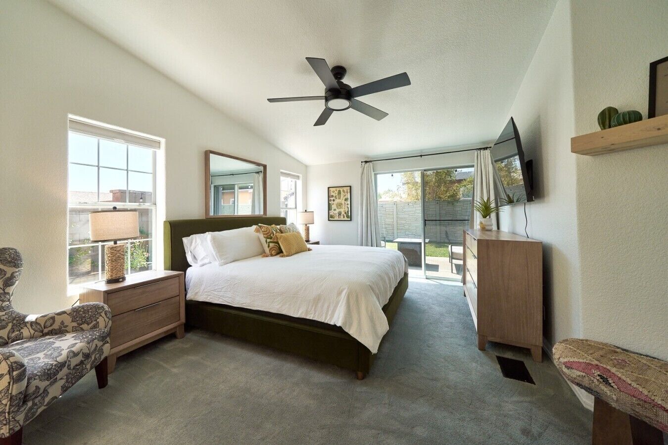 Ein Schlafzimmer mit einem Bett, einem Nachttisch, einem Sessel, einer Kommode, einem Deckenventilator und einem an der Wand montierten Fernseher.