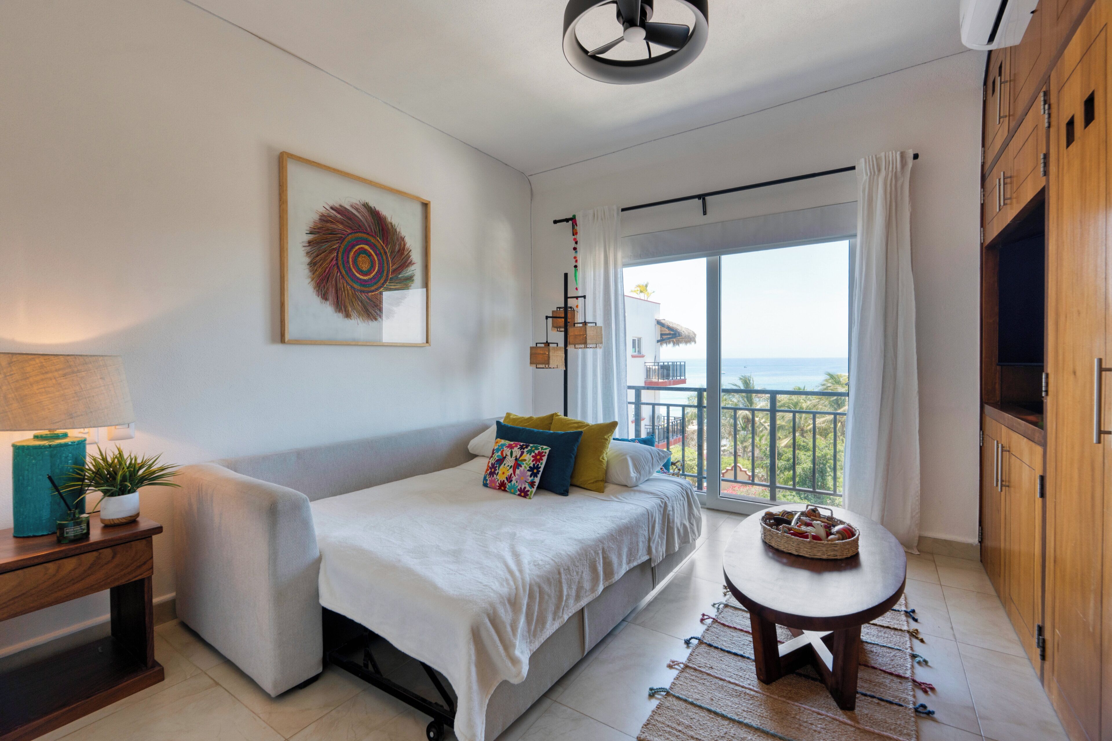 Una camera da letto moderna con balcone, un letto con molti cuscini, un tavolino con un cesto e un elemento decorativo a parete.