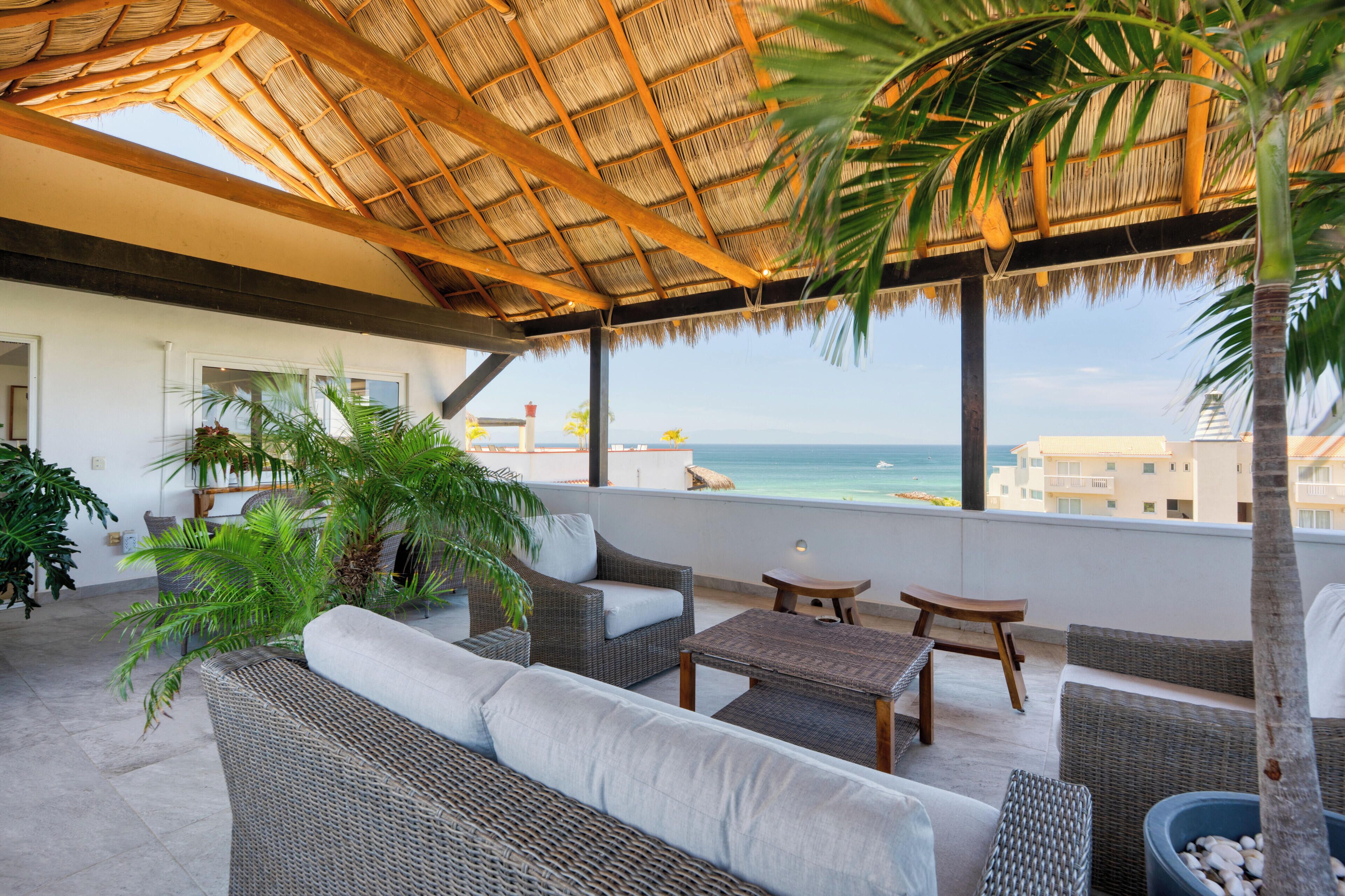 A covered outdoor seating area with wicker furniture, a thatched roof, and a view of the sea.