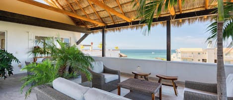 A covered outdoor seating area with wicker furniture, a thatched roof, and a view of the sea.