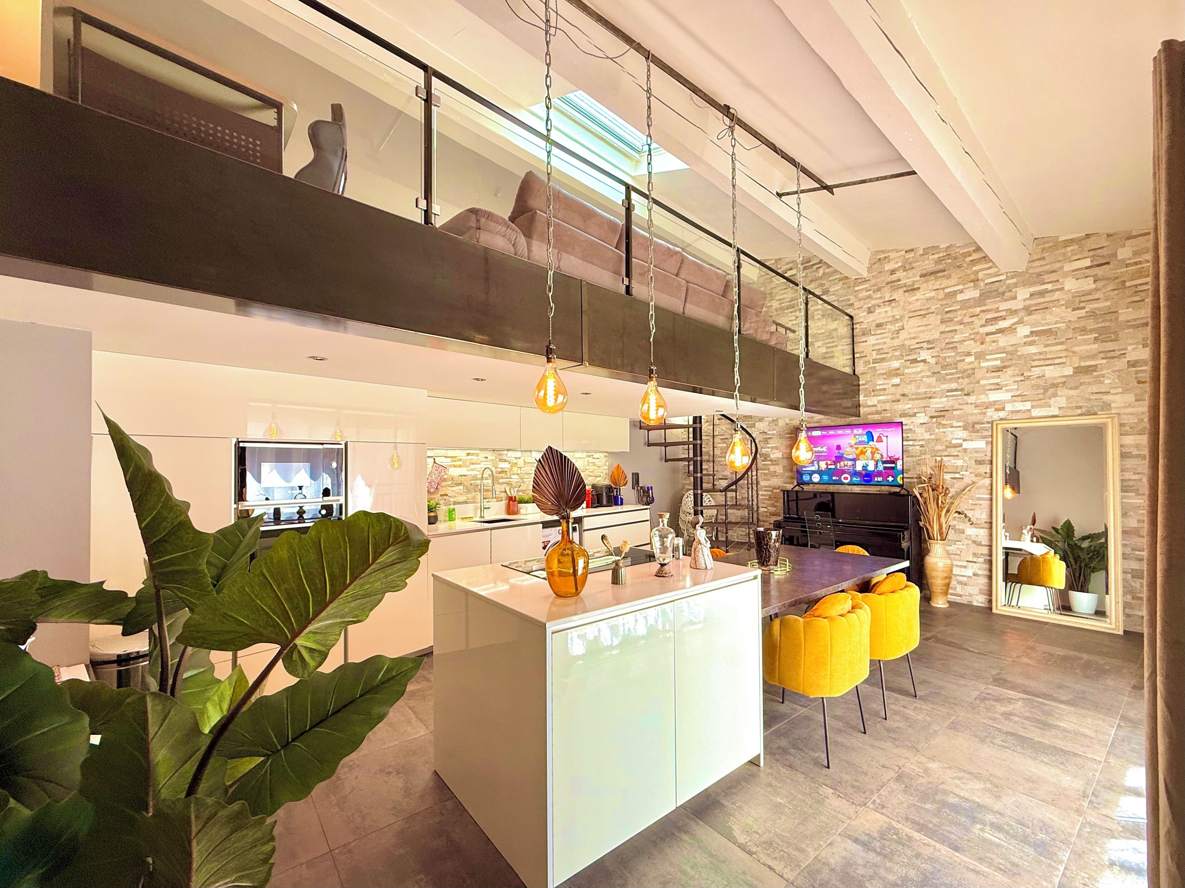 A modern kitchen with a central island, a dining area with yellow chairs, and a TV on the wall.
