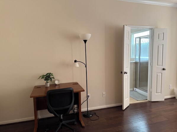 A room with a wooden desk, a black office chair, a floor lamp, a potted plant, and a white door leading to a balcony.