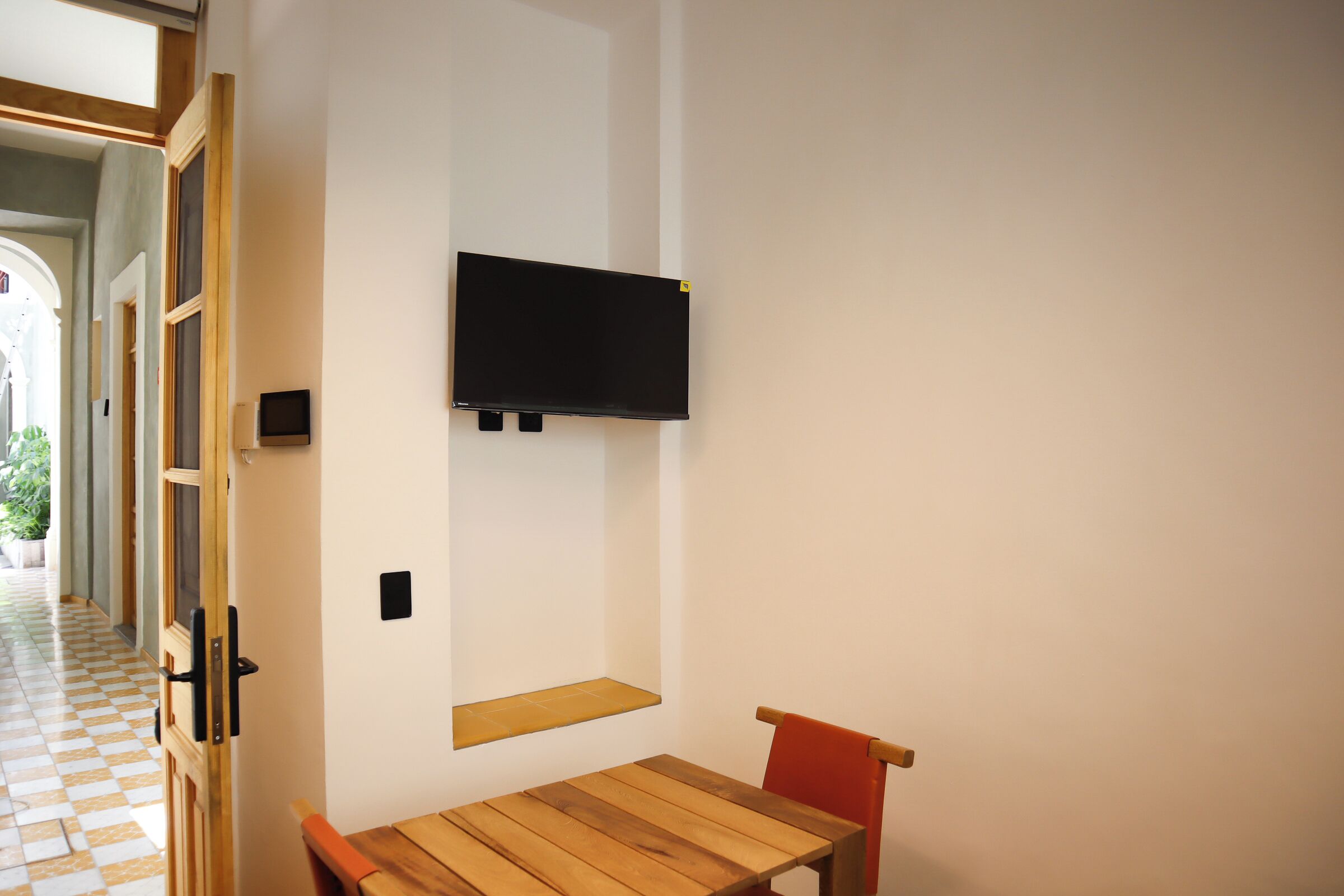 Una habitación con una mesa de madera y una silla roja, un televisor de pantalla plana montado en la pared, y un estante pequeño debajo.