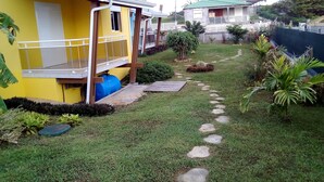 A residential area with a yellow house, a blue water tank, a paved pathway, and surrounding greenery. - BEAUVALETTE SOLEIL (Sainte-Anne)