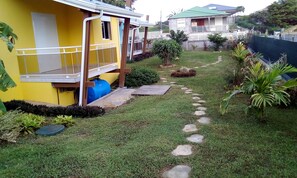 A residential area with a yellow house, a blue water tank, a paved pathway, and surrounding greenery. - BEAUVALETTE SOLEIL (Sainte-Anne)