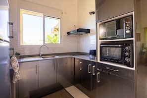 A modern kitchen with a built-in oven, microwave, and a window with a view of greenery. - BEAUVALETTE SOLEIL (Sainte-Anne)