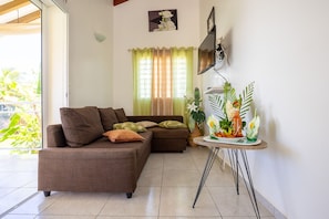 A living room with a brown sofa, a small table with a plant, and a window with curtains. - BEAUVALETTE SOLEIL (Sainte-Anne)