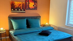 Une chambre à coucher comprenant un lit, deux œuvres d’art accrochées au mur, une table de chevet avec une lampe et une fenêtre avec des stores.