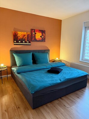 Ein Schlafzimmer mit einem Bett, zwei Wandbildern, einem Nachttisch mit Lampe und einem Fenster mit Jalousien.