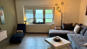 Un salon comprenant un canapé, un fauteuil, une table basse, un radiateur et une fenêtre avec des stores.