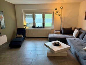Ein Wohnzimmer mit einer Couch, einem Sessel, einem Couchtisch, einer Heizung und einem Fenster mit Jalousien.