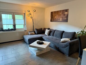 Un salon moderne avec un canapé d’angle, une table basse, un lampadaire et une décoration représentant un paysage urbain sur le rebord de la fenêtre.