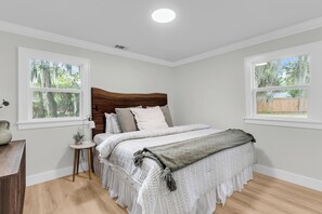Een slaapkamer met een houten hoofdeinde, een bed met witte beddengoed, twee ramen met uitzicht op bomen, een klein nachtkastje met een lamp en een houten dressoir.