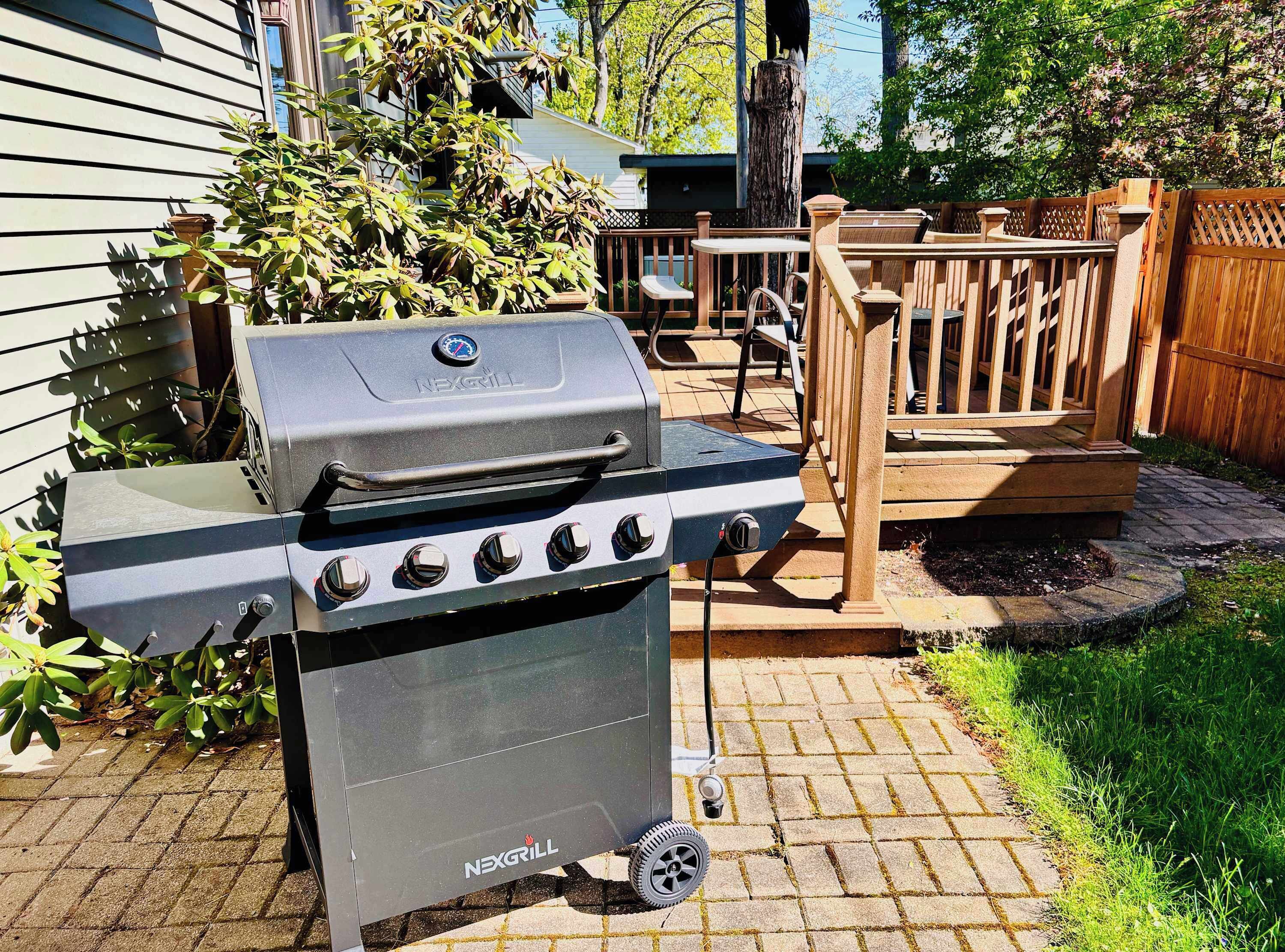 A NEXGRILL barbecue grill with four burners, a side burner, and a chimney, placed on a brick patio with a wooden deck and a wooden fence in the background.