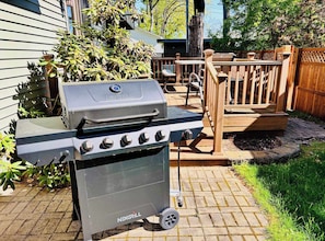 Un barbecue NEXGRILL doté de quatre brûleurs, d’un brûleur latéral et d’une cheminée, installé sur une terrasse en briques avec une plateforme en bois et une clôture en bois en arrière-plan.