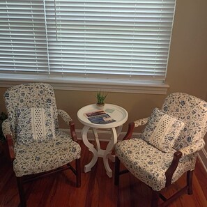Deux fauteuils à motifs, placés face à face, avec une petite table ronde entre eux, une plante en pot sur la table et, en arrière-plan, une fenêtre avec des stores.