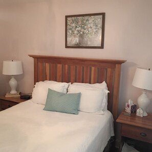 Un lit bien fait, avec une tête de lit en bois, deux lampes de chevet et une œuvre d’art florale encadrée accrochée au mur.