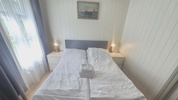 A neatly made bed with white linens, two bedside lamps, a framed picture on the wall, and a window with curtains.