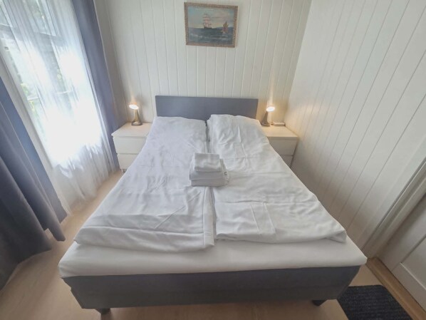A neatly made bed with white linens, two bedside lamps, a framed picture on the wall, and a window with curtains.