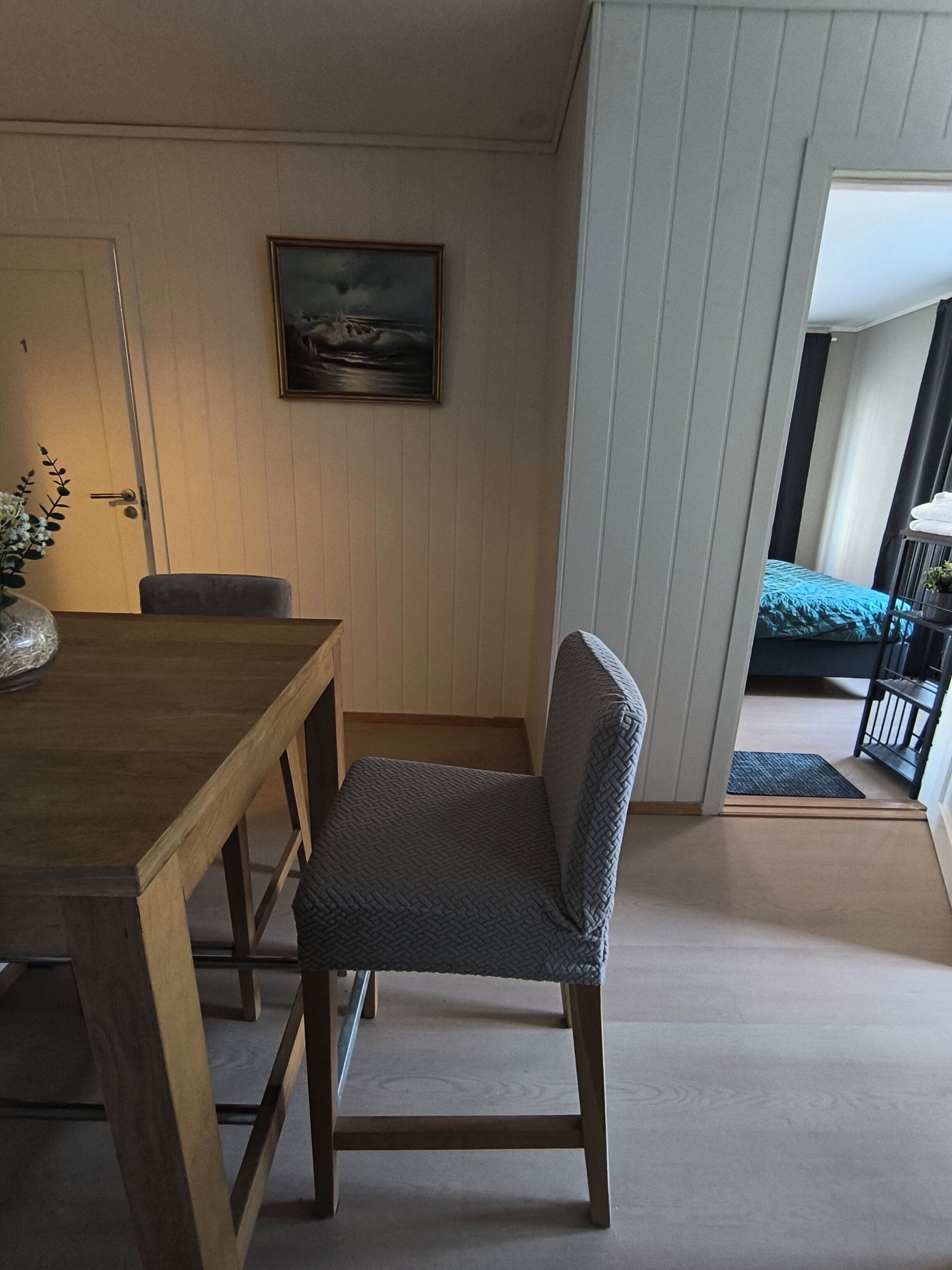 A room with a wooden dining table, a grey cushioned chair, a framed picture on the wall, and a view of a balcony with a bed.