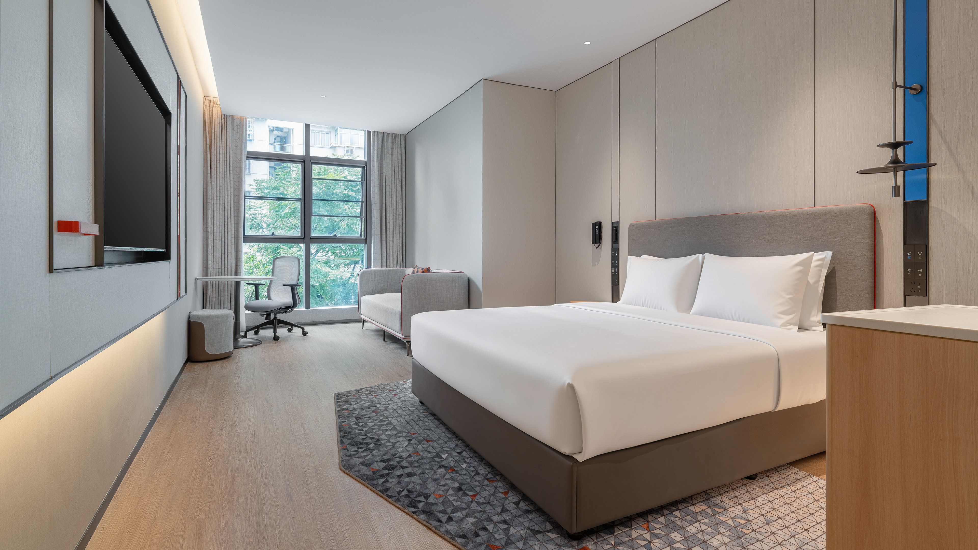 A modern hotel room with a large bed, a desk, a chair, and a view of the outdoors through the window.