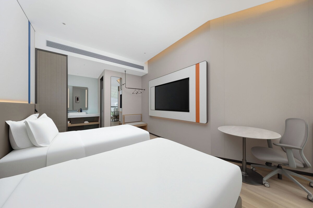 A hotel room with two beds, a TV mounted on the wall, a desk with a chair, and a bathroom visible through an open door.