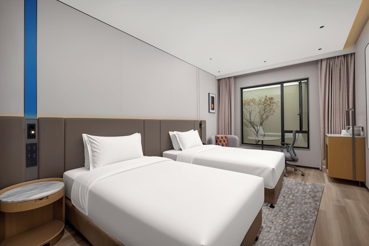 Habitación de hotel con dos camas, un escritorio, una silla, una mesita y un ventanal con cortinas.