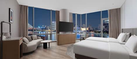 Una habitación de hotel moderna con una cama grande, un sofá, un televisor y vistas a un paisaje urbano nocturno.