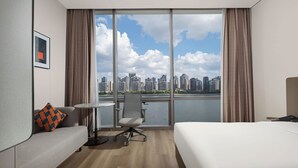 A hotel room with a bed, a sofa, a small table, and a chair. There is a large window with a view of a city skyline and a body of water. - Holiday Inn Express Shanghai North Bund Riverside By Ihg (Shanghai)