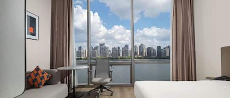 A hotel room with a bed, a sofa, a small table, and a chair. There is a large window with a view of a city skyline and a body of water.
