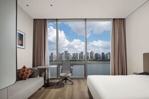 A hotel room with a bed, a sofa, a small table, and a chair. There is a large window with a view of a city skyline and a body of water. - Holiday Inn Express Shanghai North Bund Riverside By Ihg (Shanghai)