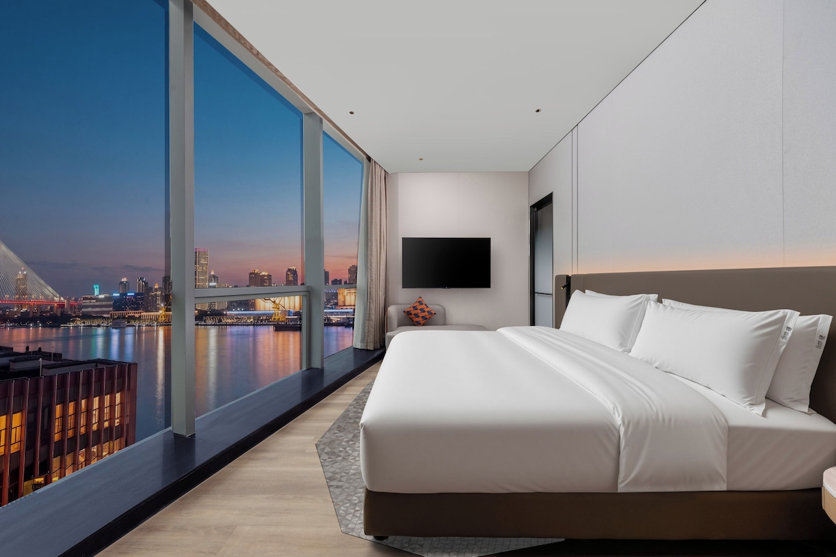 Habitación de hotel moderna con una cama grande, un televisor de pantalla plana y vistas a la silueta urbana al anochecer.
