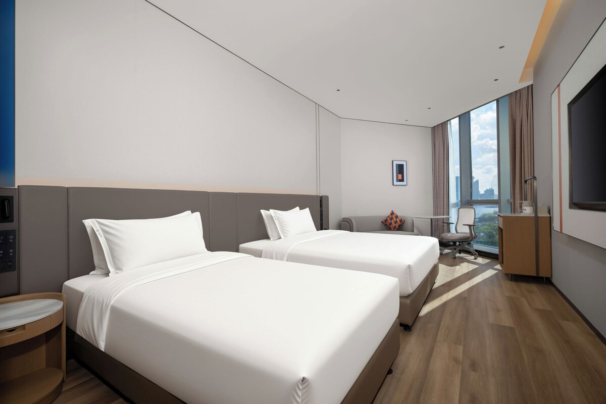 A hotel room with two beds, a desk, a chair, a TV, and a large window with a city view.
