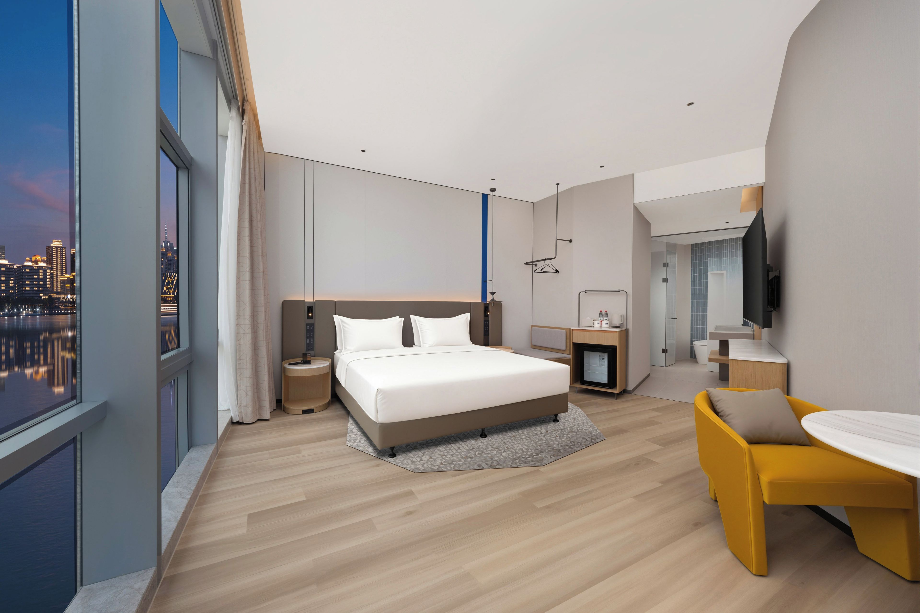 Quarto de hotel moderno com uma cama grande, uma escrivaninha, uma cadeira e vista para a paisagem urbana.