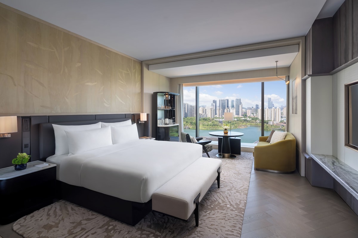 Ein modernes Hotelzimmer mit einem großen Bett, einer Sitzecke mit Stadtblick und einem kleinen Tisch mit Lampe.