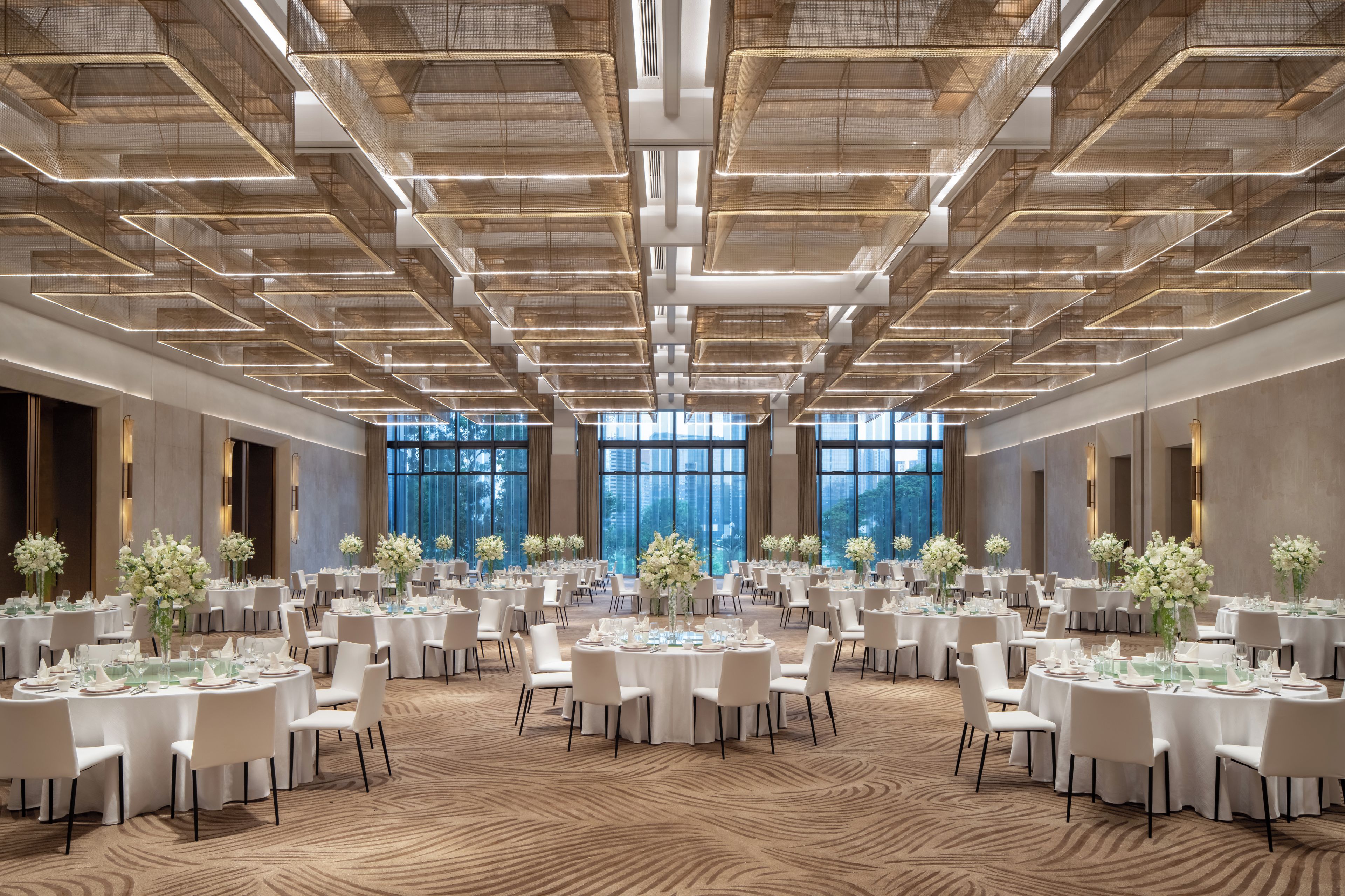 A spacious banquet hall with multiple round tables set for dining, large windows providing natural light, and a modern ceiling design.