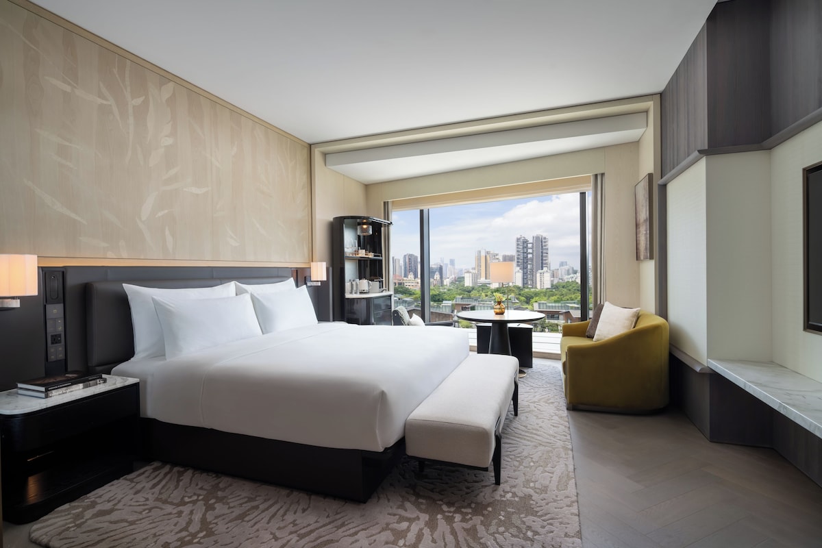 Una habitación de hotel moderna con una cama grande, una zona de estar con sofá y sillón, una mesita y vistas a la ciudad.