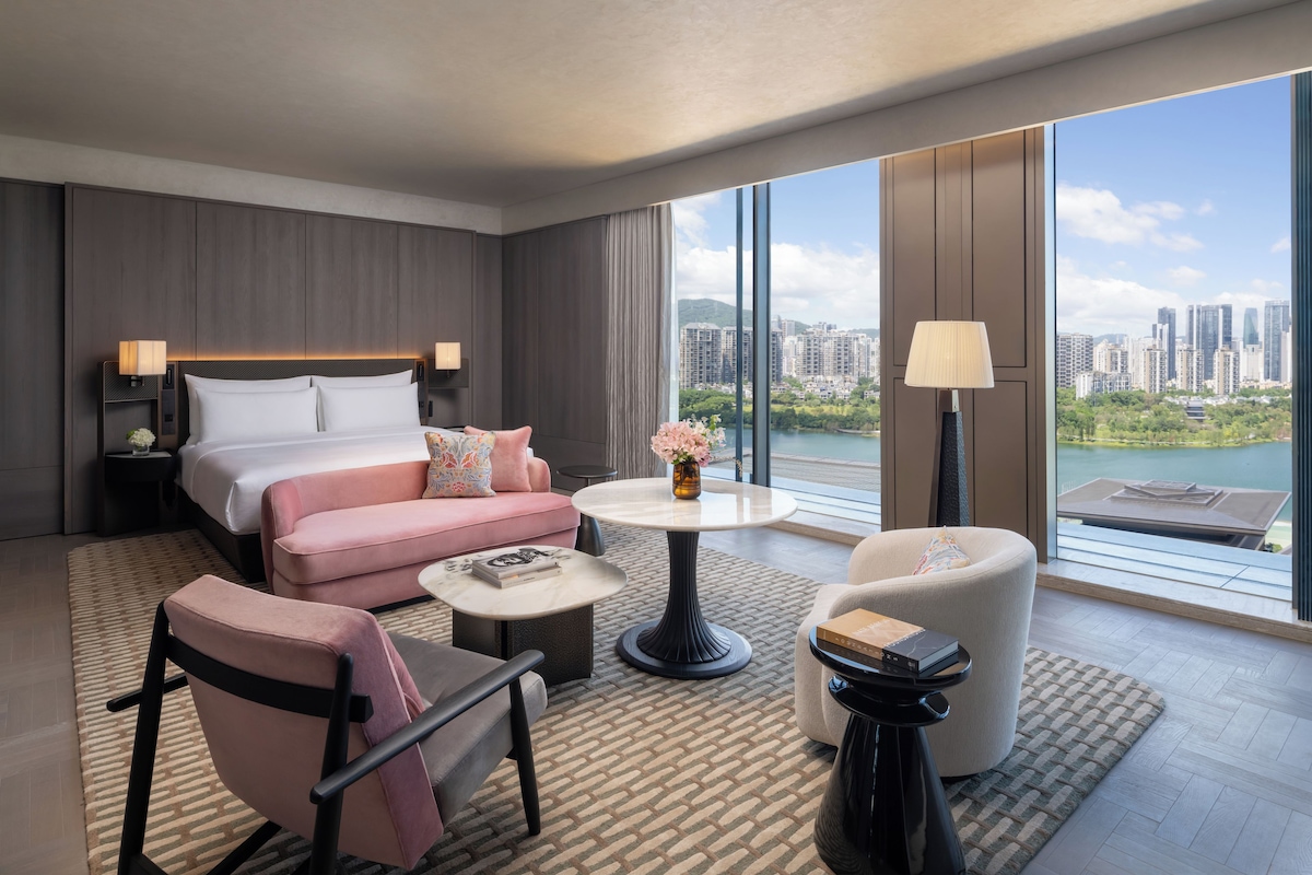 Ein modernes Hotelzimmer mit einem großen Bett, einer Couch, einem Couchtisch und einem Sessel. Durch große Fenster bietet sich ein Blick auf die Stadt.