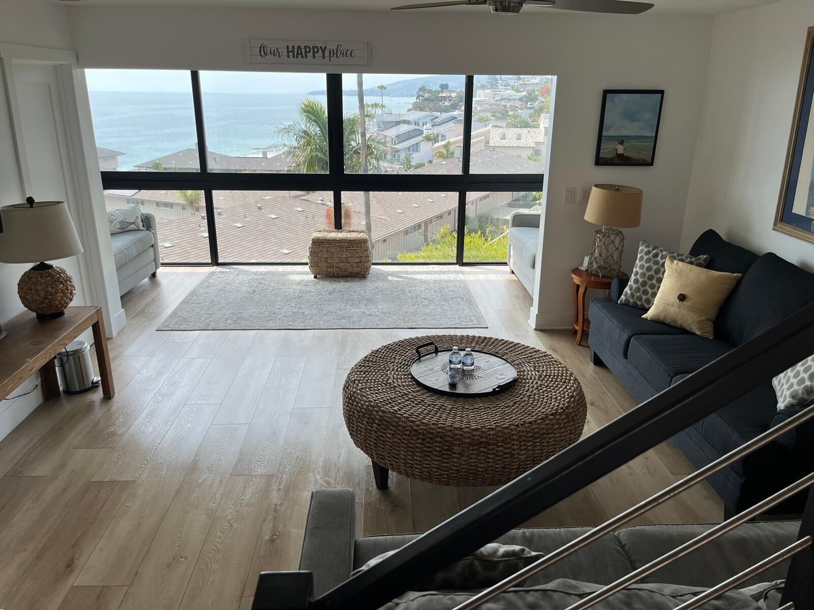 A spacious living room with a large window overlooking a coastal view, a comfortable sofa, a round wicker ottoman, and a staircase.