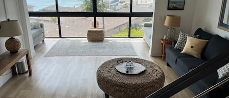 A spacious living room with a large window overlooking a coastal view, a comfortable sofa, a round wicker ottoman, and a staircase.