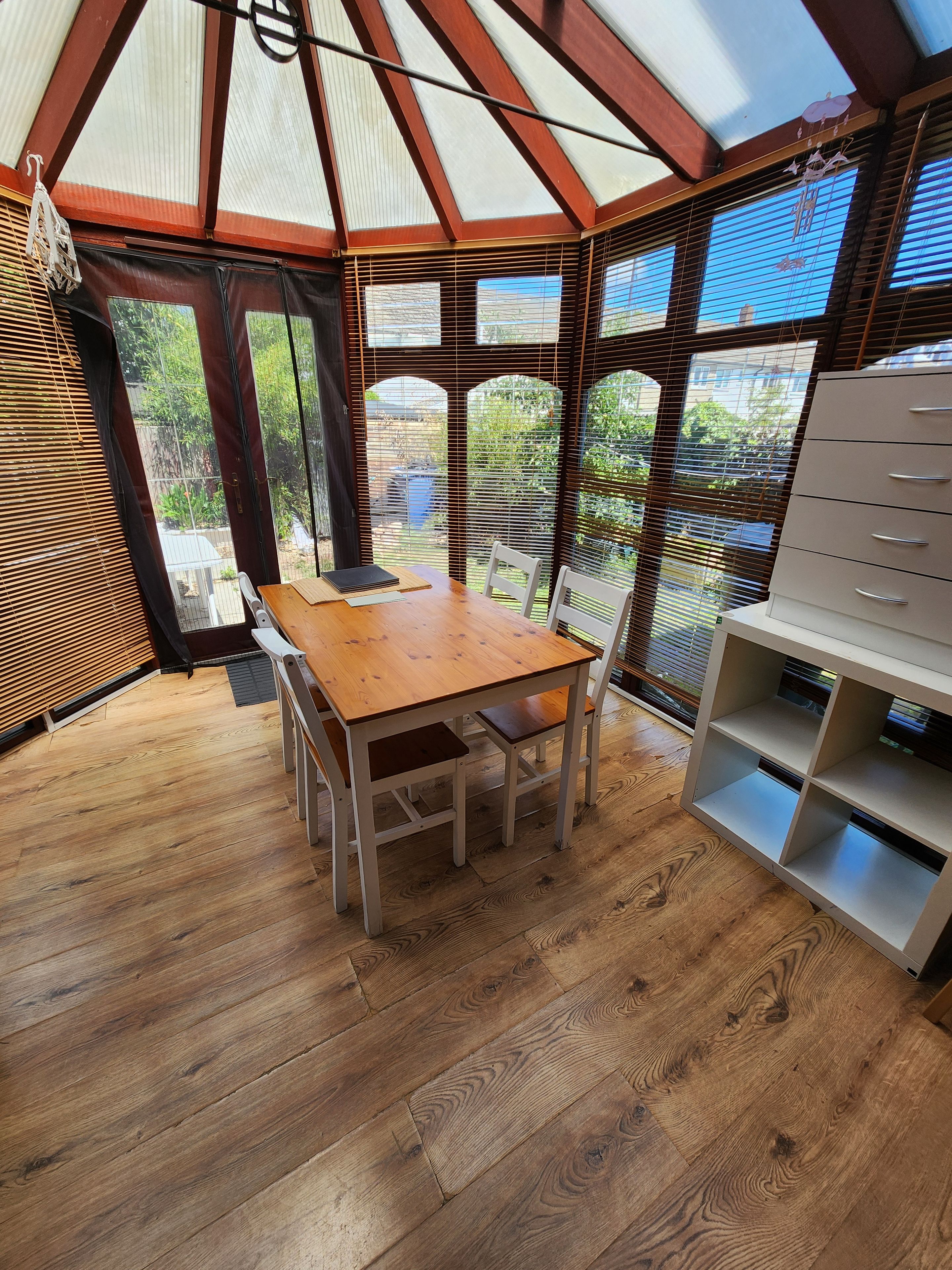 A conservatory with a wooden table and chairs, a white shelving unit, and a view of greenery outside.